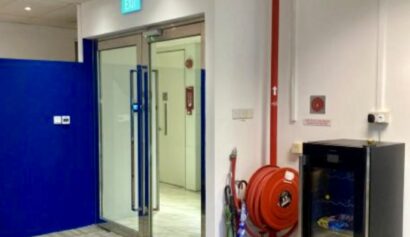 Office corridor with a glass door exit, fire hose reel on the wall, umbrella stand, and a small glass-fronted fridge.