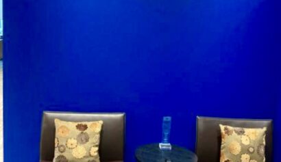 Two brown chairs with floral cushions are placed beside a small round table holding a blue award, all set against a bright blue wall in an office space.