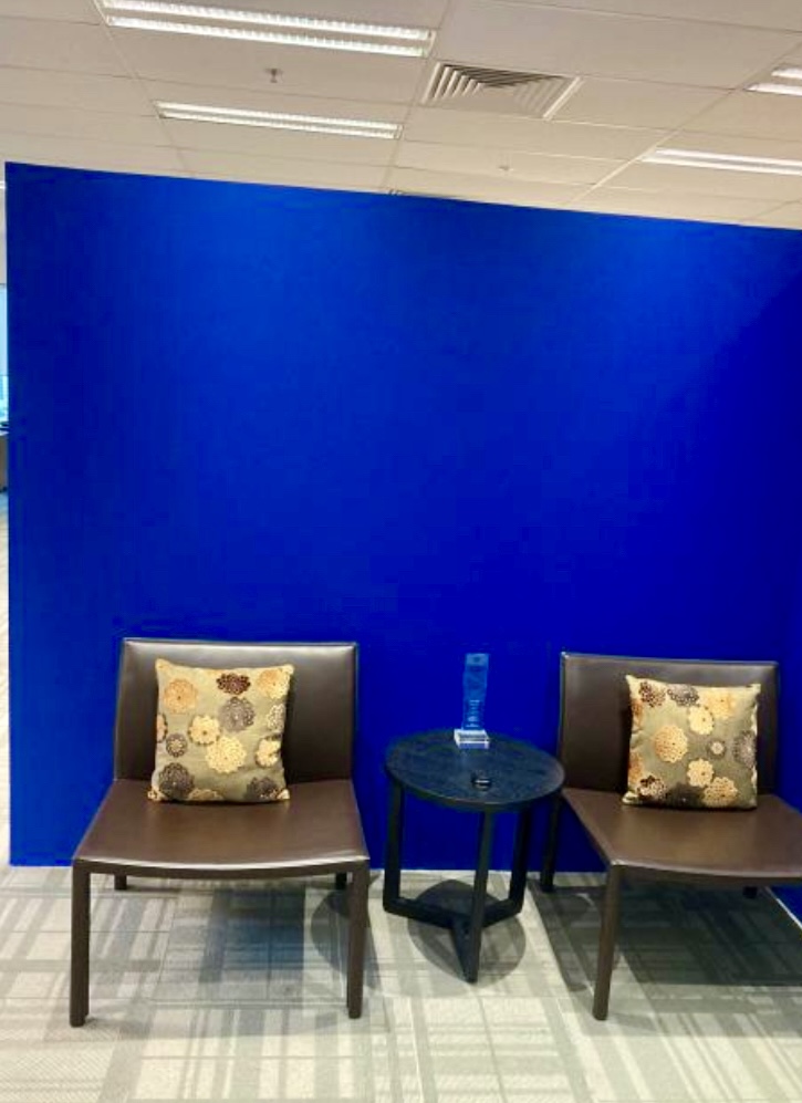 Two brown chairs with floral cushions are placed beside a small round table holding a blue award, all set against a bright blue wall in an office space.