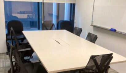 A small conference room with a rectangular table, six black office chairs, a whiteboard on the wall, and windows with blinds partially pulled down.