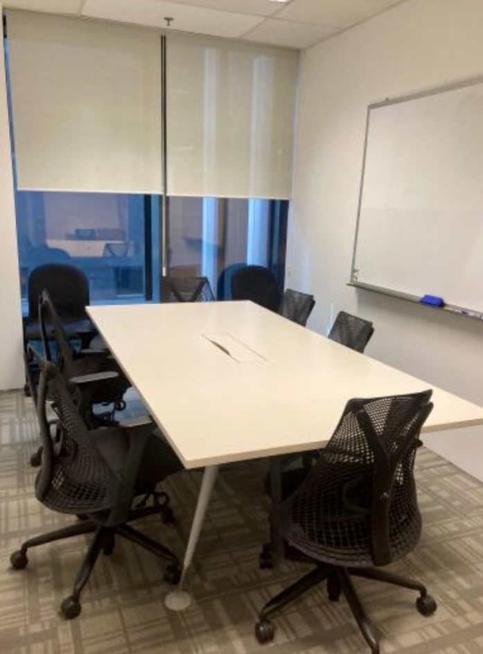 A small conference room with a rectangular table, six black office chairs, a whiteboard on the wall, and windows with blinds partially pulled down.