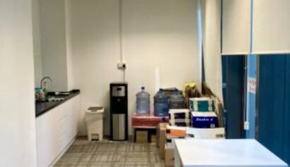 Office pantry area with a white table and chair, water dispenser, water bottles, cardboard boxes, worktop with sink, and blue blinds covering the windows.