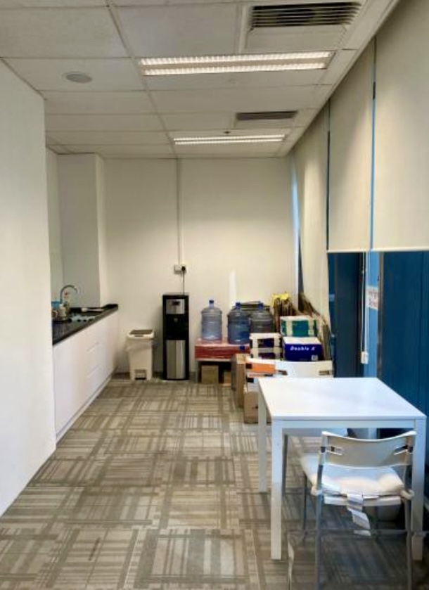 Office pantry area with a white table and chair, water dispenser, water bottles, cardboard boxes, worktop with sink, and blue blinds covering the windows.
