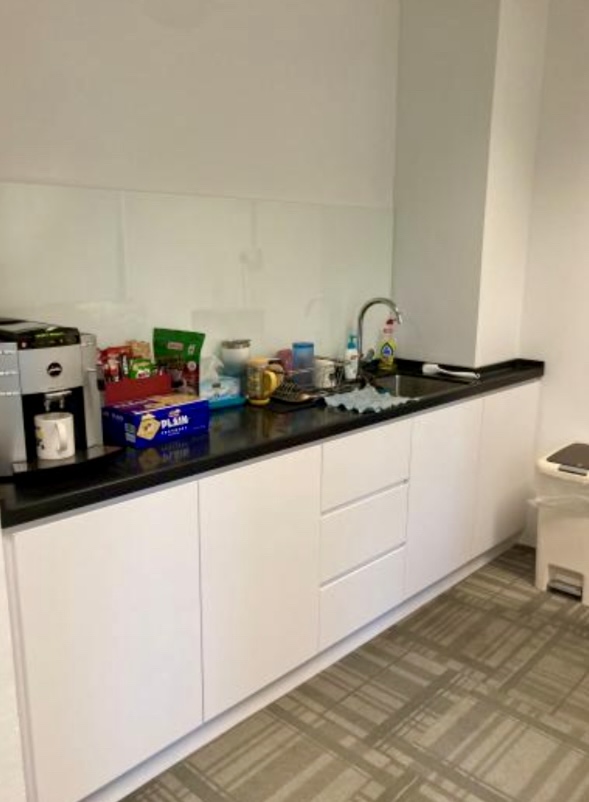 A kitchenette with white cupboards, a black worktop, various kitchen items, a coffee machine, mugs, snacks, and a sink; a rubbish bin is visible to the right.