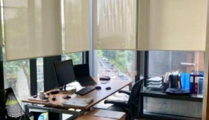 A small office with two chairs, a desk with computers and paperwork, large windows with blinds, and shelves holding files and folders.