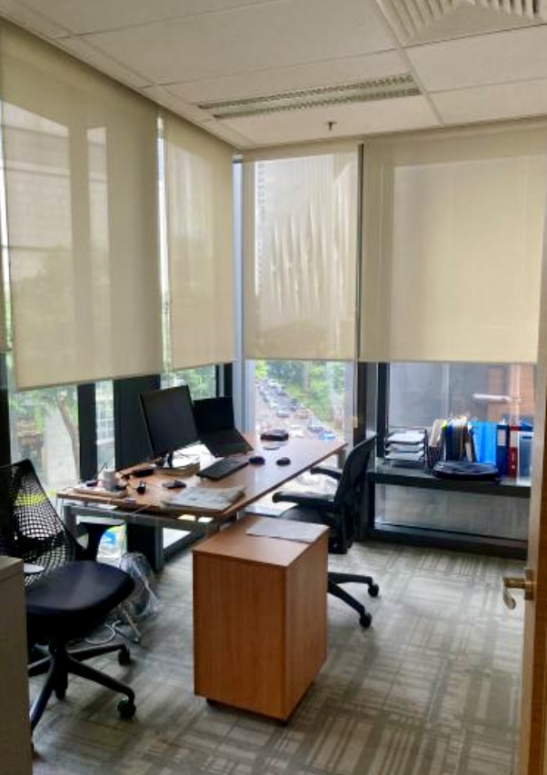 A small office with two chairs, a desk with computers and paperwork, large windows with blinds, and shelves holding files and folders.