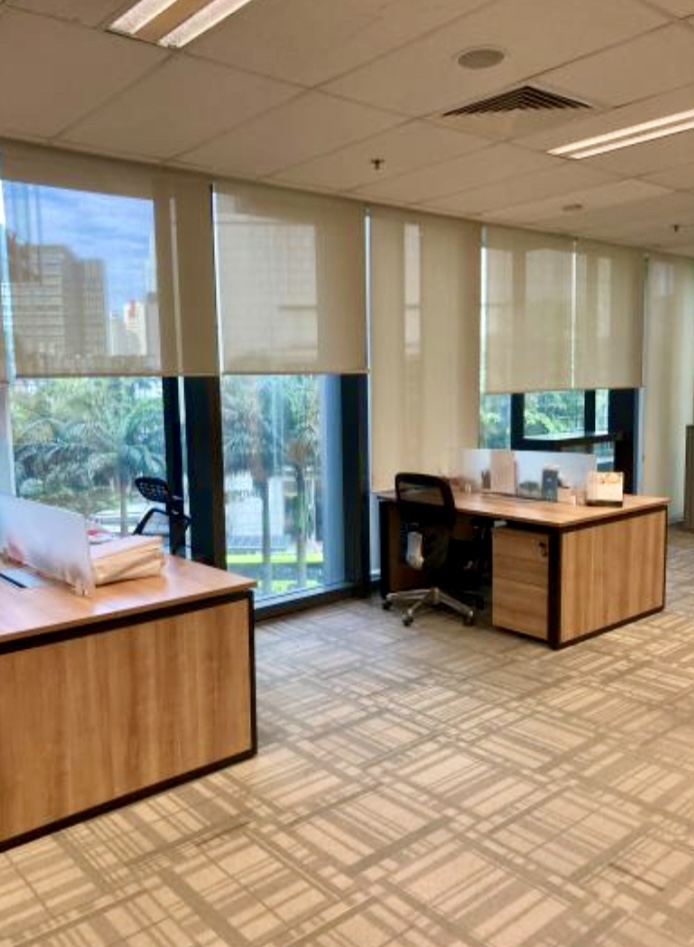 Modern office with wooden desks, black chairs, large windows with roller blinds, and a view of palm trees outside.