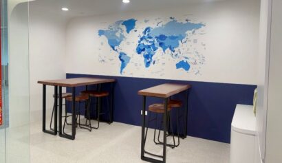 Two high tables with wooden stools are set against a white and blue wall featuring a blue world map mural in a modern office space.