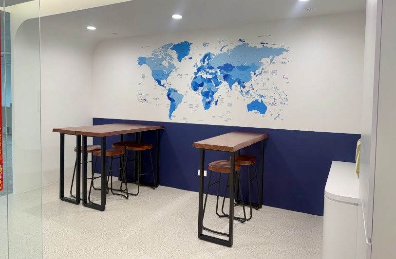 Two high tables with wooden stools are set against a white and blue wall featuring a blue world map mural in a modern office space.