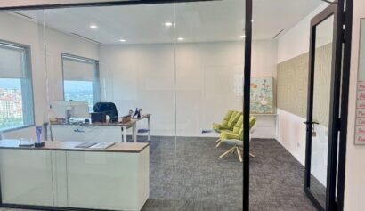 A modern glass-walled office with a desk, computer, office chair, and two green lounge chairs; large windows provide natural light.