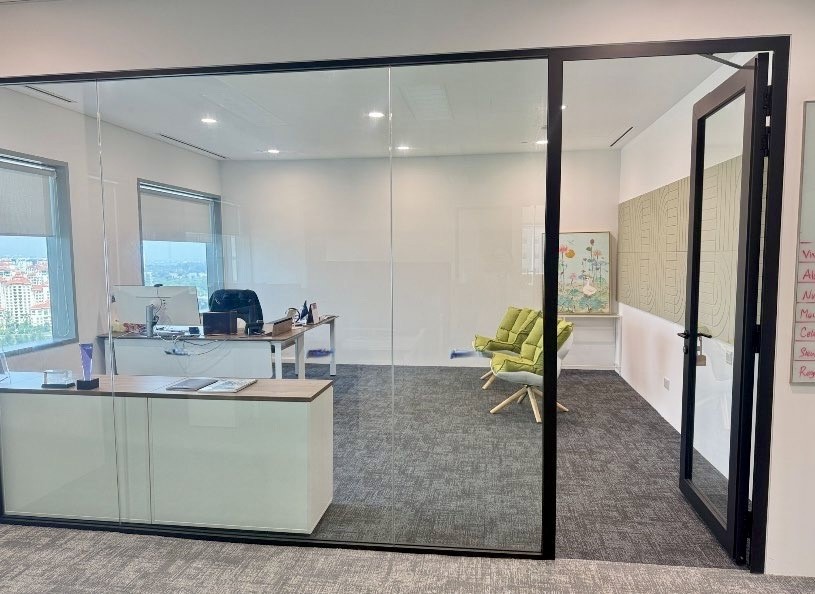 A modern glass-walled office with a desk, computer, office chair, and two green lounge chairs; large windows provide natural light.