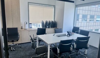A modern office meeting room with a white table, six black chairs, a conference phone, and windows overlooking another building. Five rucksacks are stacked in the corner.