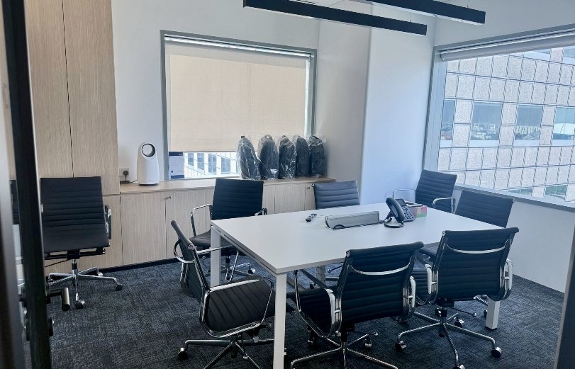 A modern office meeting room with a white table, six black chairs, a conference phone, and windows overlooking another building. Five rucksacks are stacked in the corner.