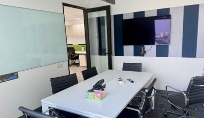 A modern conference room with a white table, six black chairs, a whiteboard, a wall-mounted TV, and an open door leading to an office area.