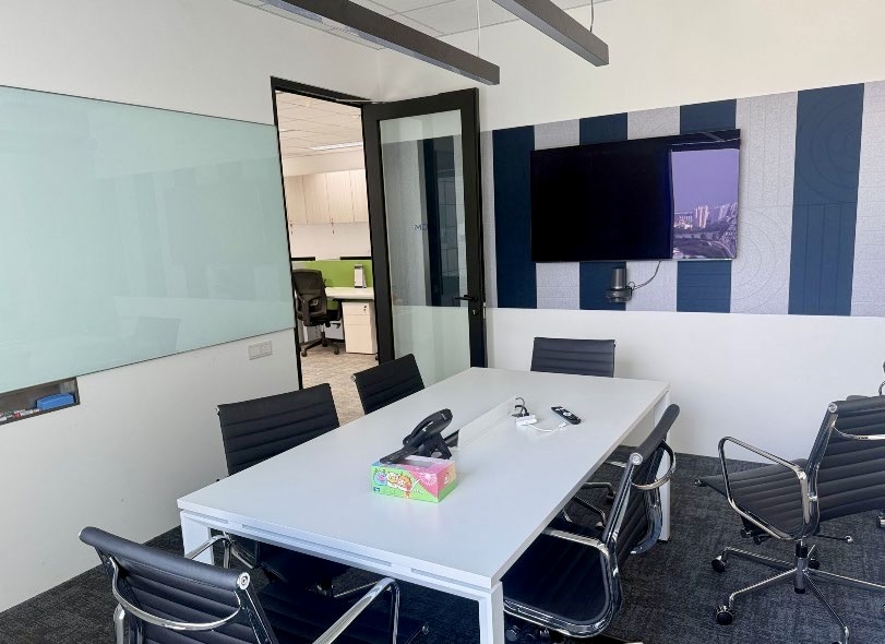 A modern conference room with a white table, six black chairs, a whiteboard, a wall-mounted TV, and an open door leading to an office area.