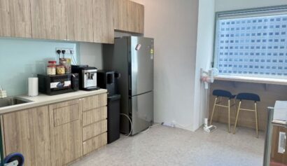 Small office pantry with wooden cupboards, a fridge, coffee machine, snacks, sink, and a worktop with two bar stools by the window overlooking tall buildings.