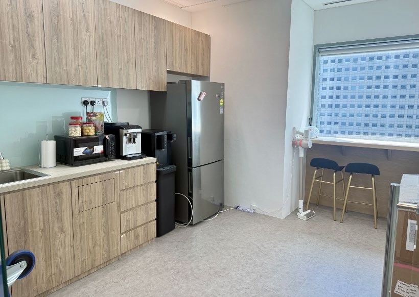 Small office pantry with wooden cupboards, a fridge, coffee machine, snacks, sink, and a worktop with two bar stools by the window overlooking tall buildings.
