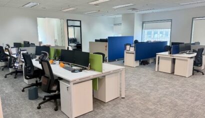 Modern open-plan office with several empty desks, chairs, desktop computers, partition screens, and large windows allowing natural light.