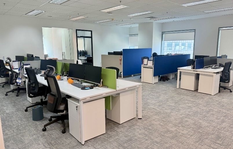 Modern open-plan office with several empty desks, chairs, desktop computers, partition screens, and large windows allowing natural light.