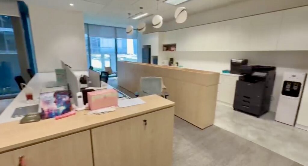 Modern office space with wood-finished desks, computers, a photocopier, water dispenser, and shelves, illuminated by natural light from large windows.