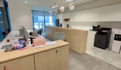 Modern office space with wood-finished desks, computers, a photocopier, water dispenser, and shelves, illuminated by natural light from large windows.