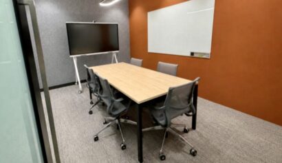 A modern conference room with a rectangular table, six grey chairs, a large screen on a stand, a whiteboard, and an orange feature wall.