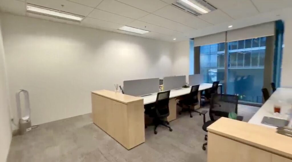 Modern office space with several desks and chairs, light wood furniture, neutral walls, large window, and natural light.