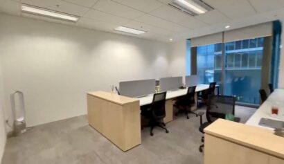 Modern office space with several desks and chairs, light wood furniture, neutral walls, large window, and natural light.