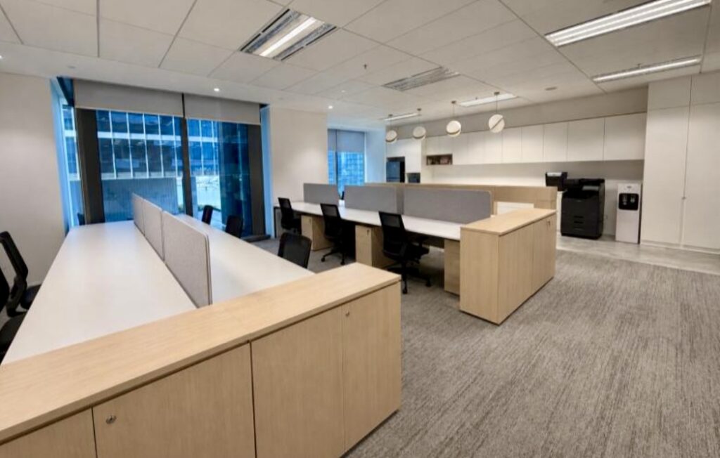 Modern open-plan office with light wood desks, black chairs, grey carpet, large windows, overhead lighting, and a photocopier in the corner.