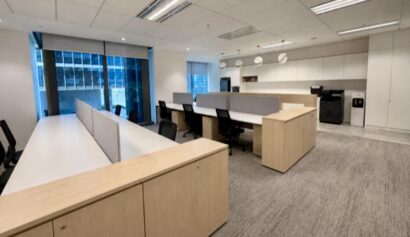 Modern open-plan office with light wood desks, black chairs, grey carpet, large windows, overhead lighting, and a photocopier in the corner.