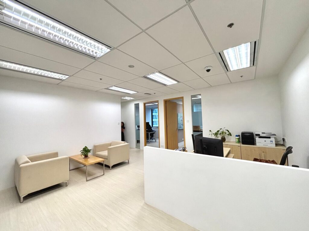 Modern office with white walls, beige chairs around a small table, a desk with a computer, printer, and a plant, and fluorescent ceiling lights.