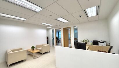 Modern office with white walls, beige chairs around a small table, a desk with a computer, printer, and a plant, and fluorescent ceiling lights.