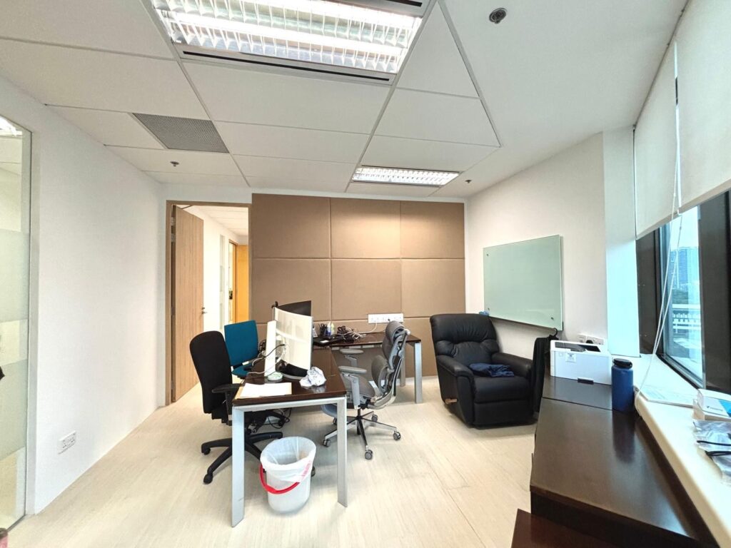 A modern office with two desks, computer monitors, office chairs, a black recliner, whiteboard, windows, and various office supplies.