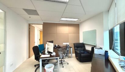A modern office with two desks, computer monitors, office chairs, a black recliner, whiteboard, windows, and various office supplies.