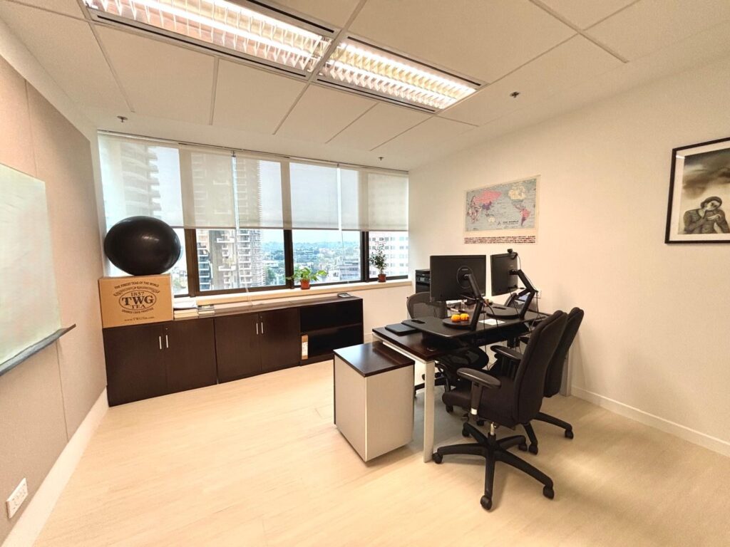 Modern office with two chairs, a desk with computer monitors, large windows with city view, a filing cabinet, and wall decorations including a map and a framed photograph.