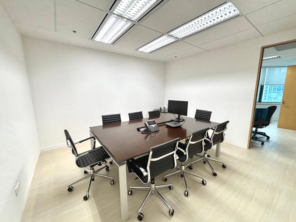 Small meeting room with a rectangular table, six black chairs, a computer monitor, a telephone, and office supplies; white walls and fluorescent ceiling lights.