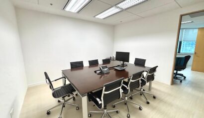 Small meeting room with a rectangular table, six black chairs, a computer monitor, a telephone, and office supplies; white walls and fluorescent ceiling lights.
