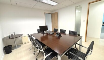 A modern conference room with a large wooden table, eight black chairs, a computer monitor, conference telephone, and minimal decor.