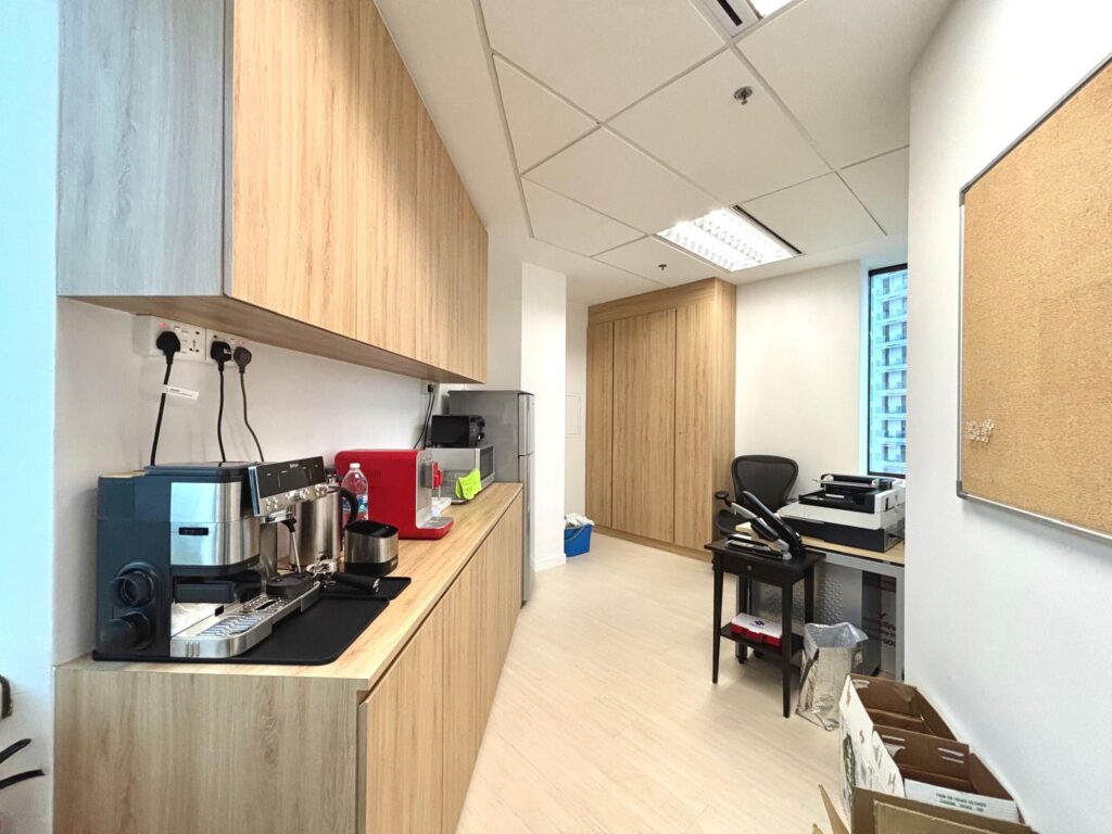 Office pantry with wooden cupboards, coffee machines, water dispenser, and a work desk with a chair; noticeboard and some boxes on the right side.