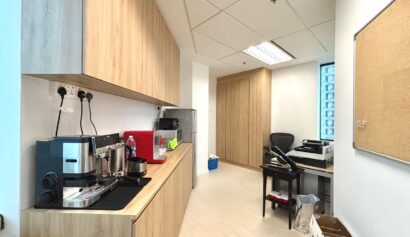 Office pantry with wooden cupboards, coffee machines, water dispenser, and a work desk with a chair; noticeboard and some boxes on the right side.
