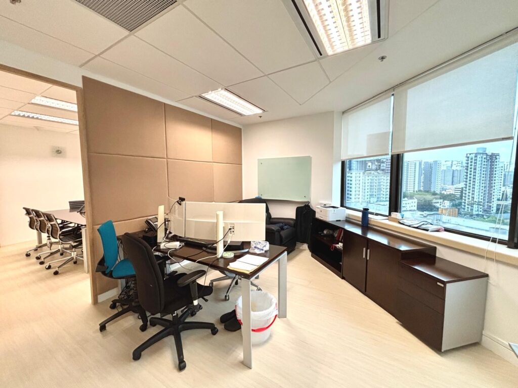 Modern office with desks, chairs, computers, whiteboard, cupboards, and large windows overlooking city buildings. Meeting room visible through open doorway.