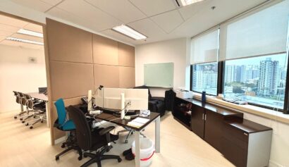 Modern office with desks, chairs, computers, whiteboard, cupboards, and large windows overlooking city buildings. Meeting room visible through open doorway.