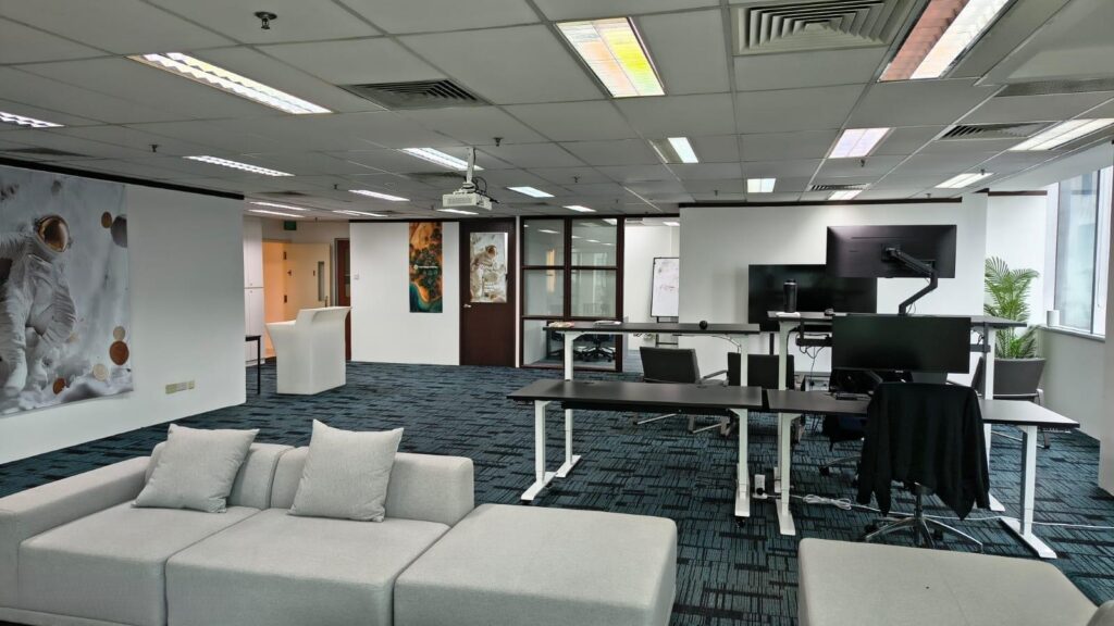 Modern office with desks, computers, a grey sofa, wall art featuring an astronaut, large windows, and a whiteboard in the background.