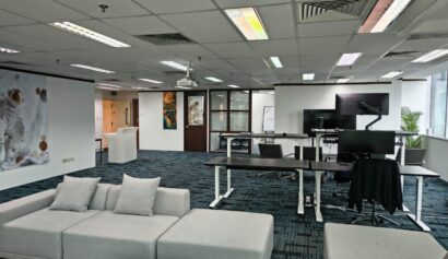 Modern office with desks, computers, a grey sofa, wall art featuring an astronaut, large windows, and a whiteboard in the background.