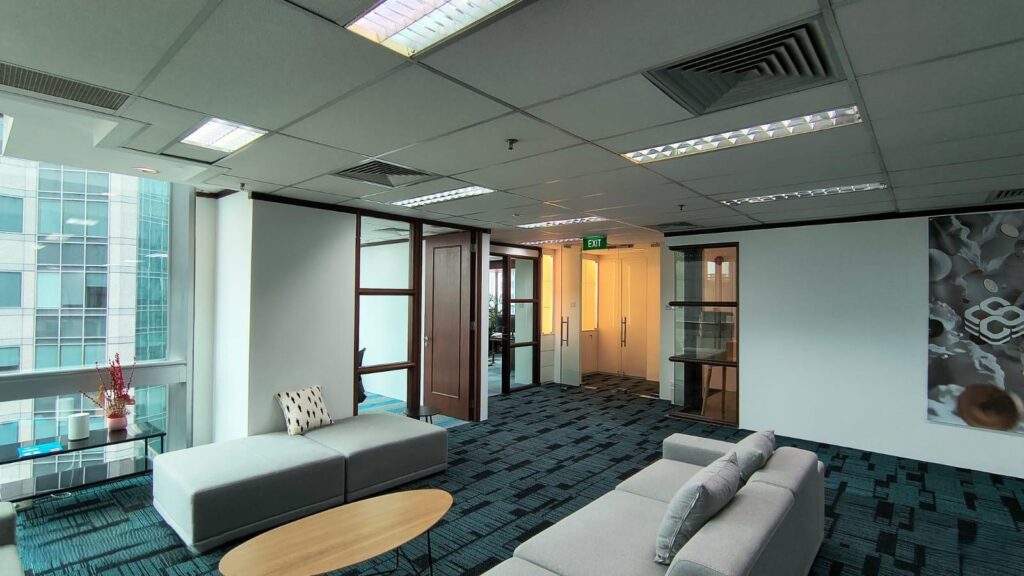 Modern office lounge with grey sofas, a round wooden coffee table, blue carpet, glass doors, and large windows overlooking adjacent buildings.