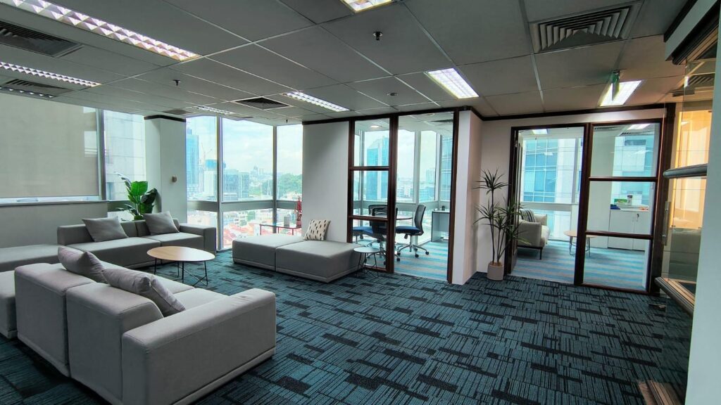 Modern office space with large windows, grey sofas, round tables, glass-walled meeting rooms, and a potted plant on blue-patterned carpet.