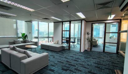Modern office space with large windows, grey sofas, round tables, glass-walled meeting rooms, and a potted plant on blue-patterned carpet.
