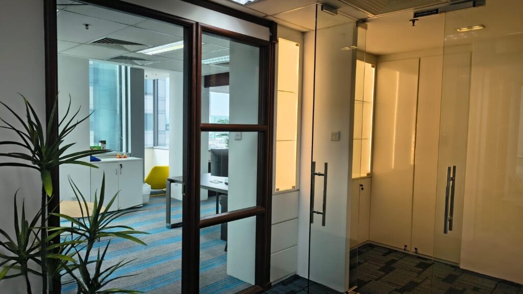 A modern office space with glass doors, a potted plant, blue carpet, a small table with a plant, and large windows letting in natural light.