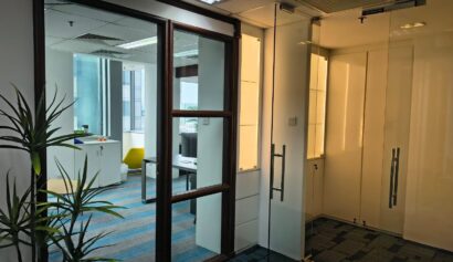 A modern office space with glass doors, a potted plant, blue carpet, a small table with a plant, and large windows letting in natural light.