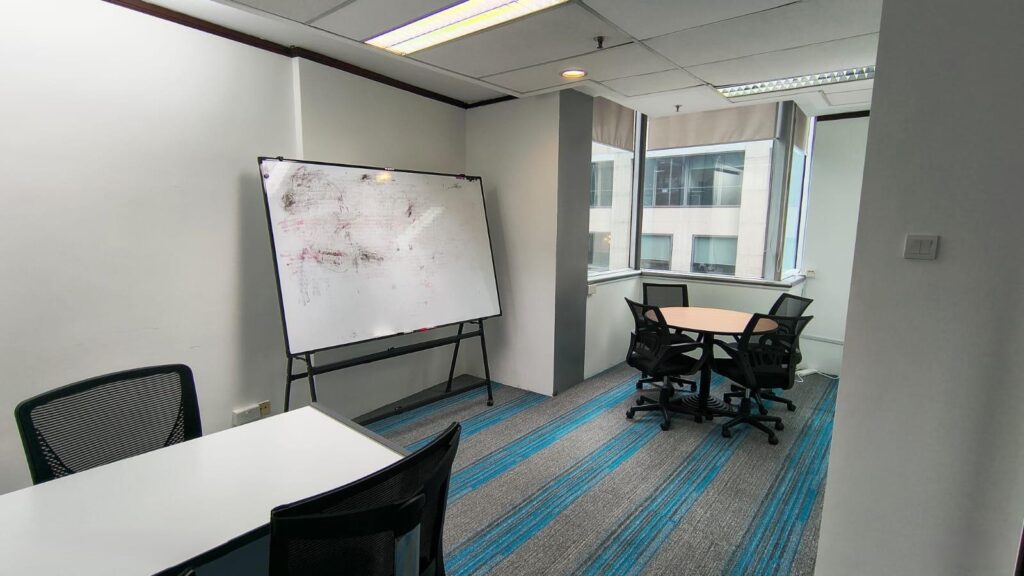 A small office room with a whiteboard, two tables, several black chairs, and large windows letting in natural light.